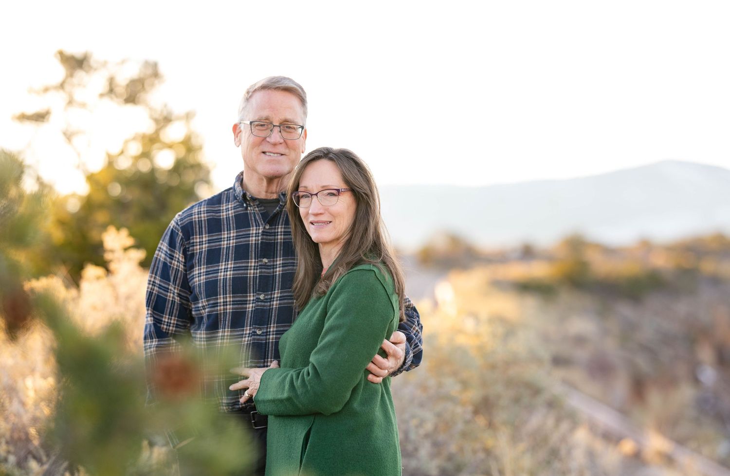 An affectionate couple poses outdoors at golden hour in a desert landscape with mountains in the background.