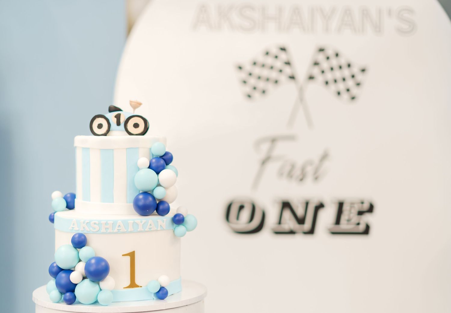 A white and blue birthday cake decorated with balloons and a race car topper for a first birthday celebration.