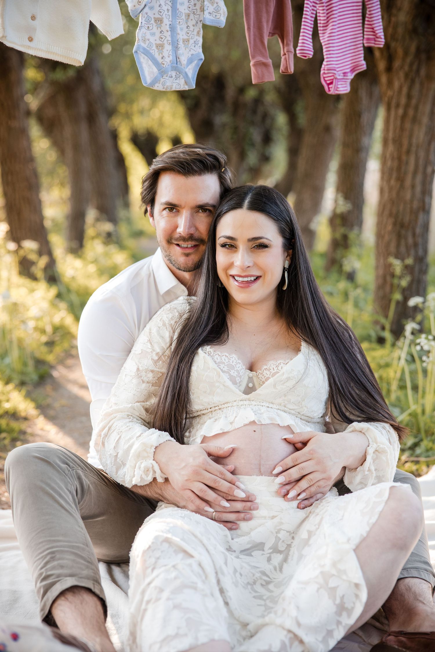A couple in white and beige clothing pose together outdoors for maternity photos in a natural setting.