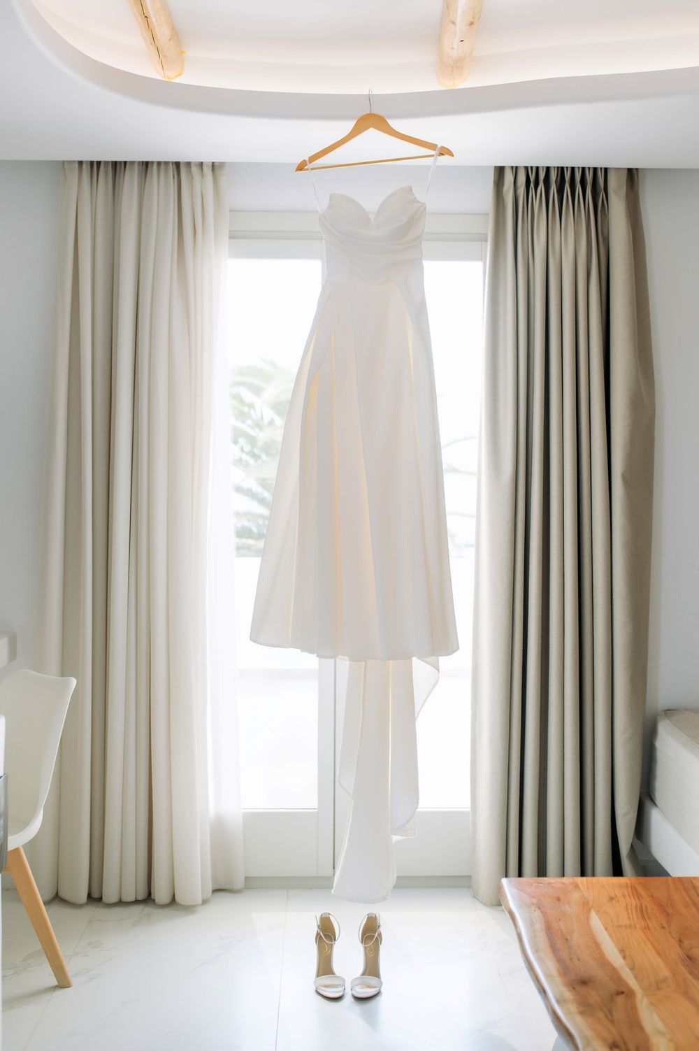 A white wedding dress and heels hanging in a bright room with neutral curtains.