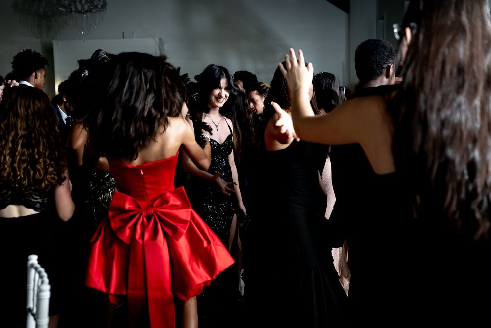 A crowd dances at a party while someone in a bright red dress with a bow stands out among others in black attire.
