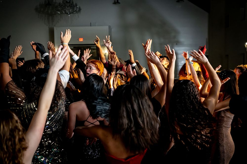 Enthusiastic crowd of concert attendees raise their hands in unison during a lively indoor performance.