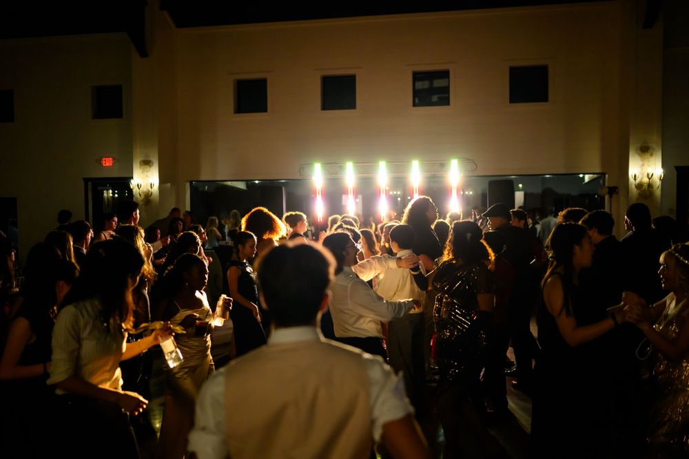 A crowded dance floor of party guests illuminated by stage lights in a dimly lit reception hall.