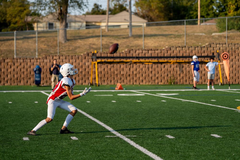 A lacrosse player in white and red uniform practices on an artificial turf field during a sunny afternoon.