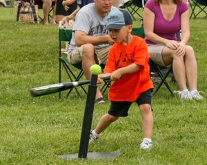 Young child in orange shirt plays tee ball on a grassy field while spectators watch from folding chairs.