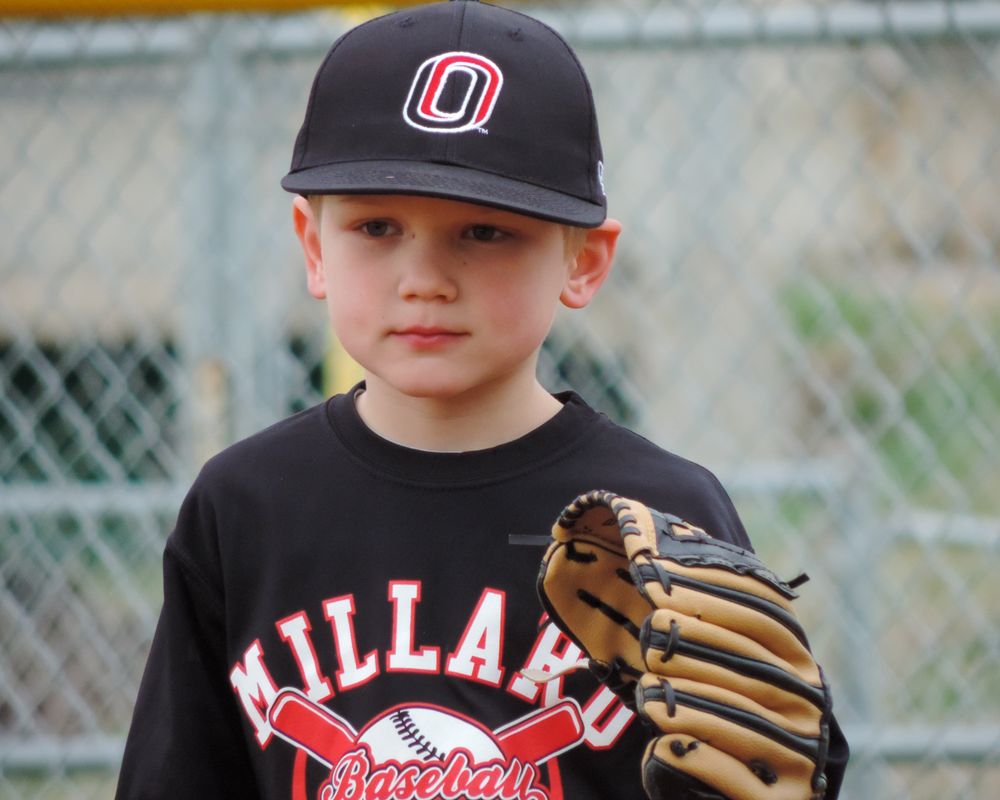 Young baseball player wearing a black Millard Dawgs uniform and cap poses with baseball glove.