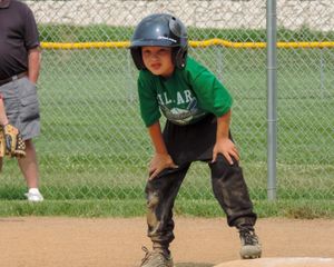 Young baseball player in green jersey and helmet takes a lead off base during a game.