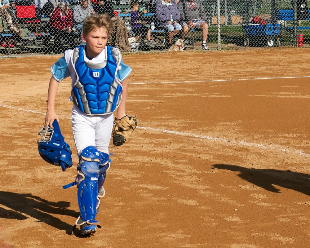 Baseball catcher in blue gear crouches on a dusty diamond field with spectators visible in the background.