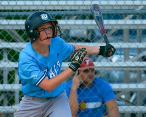 Youth baseball batter in light blue jersey stands ready at home plate with metal bat during game.