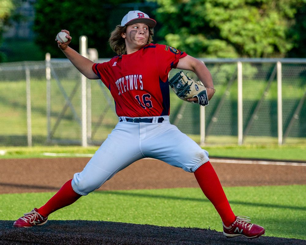 Baseball pitcher in red jersey and gray pants mid-delivery on mound during game action.