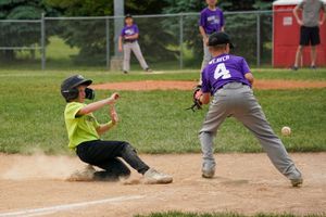 Baseball player in yellow jersey slides into home plate as fielder in purple uniform tries to make the play.