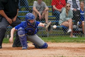 Baseball catcher in blue uniform crouches to catch a ball during game with spectators behind fence in background.