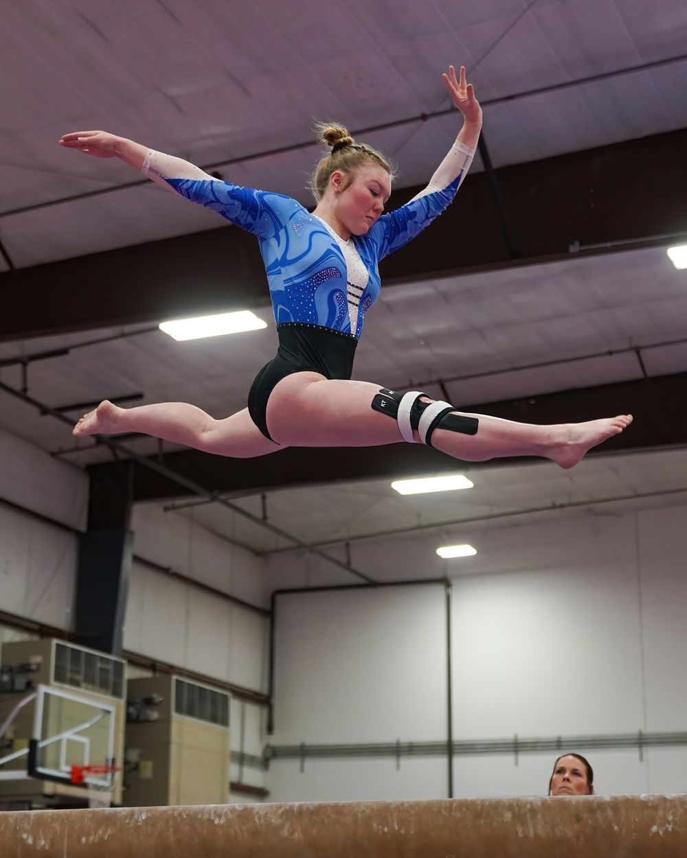 A gymnast in a blue leotard jumps high with a split leap over a balance beam in an indoor gymnasium.