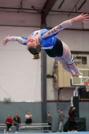 Gymnast in blue leotard performs a back layout during competition with spectators watching in the background.
