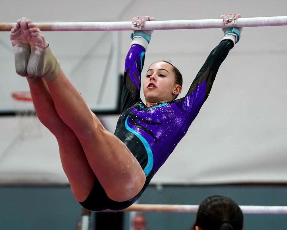 A gymnast in a purple and turquoise leotard performs on the uneven bars during competition.
