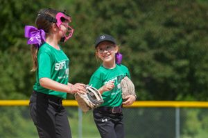 Two softball players in matching green jerseys and purple hair bows share a laugh during a game on the field.