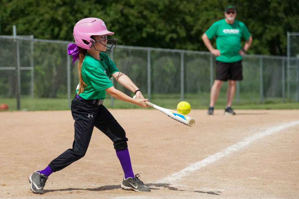 Youth softball player in green jersey and pink helmet swings at a pitch during a game on a dirt field.