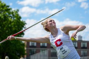 Athlete in white uniform throwing javelin during track and field competition with blue sky and trees in background.