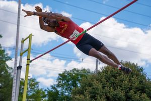 Athlete in red jersey performing high jump at outdoor track meet against blue sky with clouds.