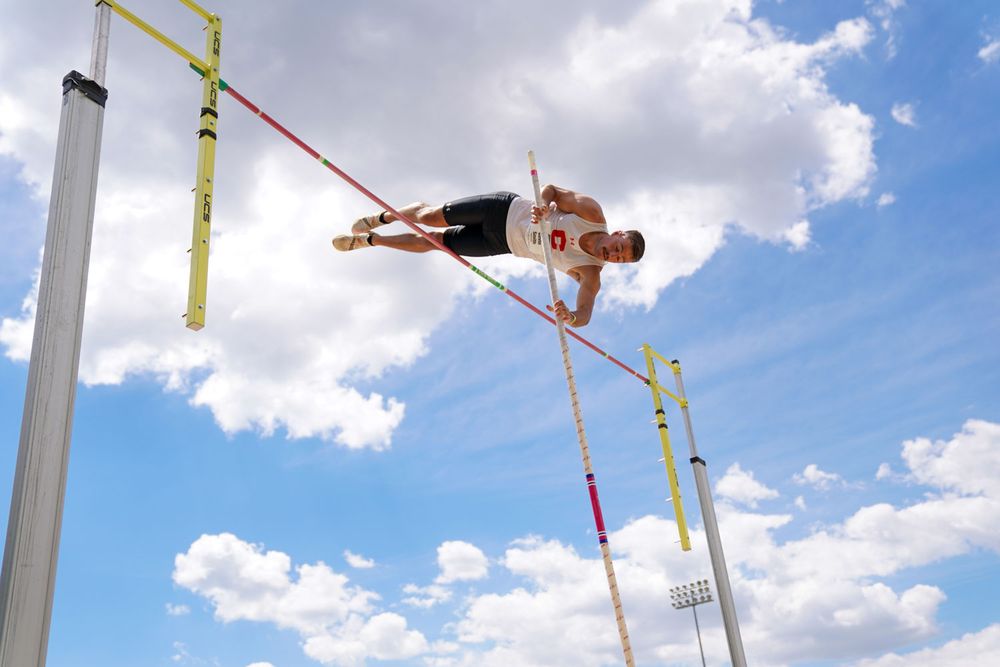 Athlete performing a pole vault jump against a bright blue sky with white clouds.