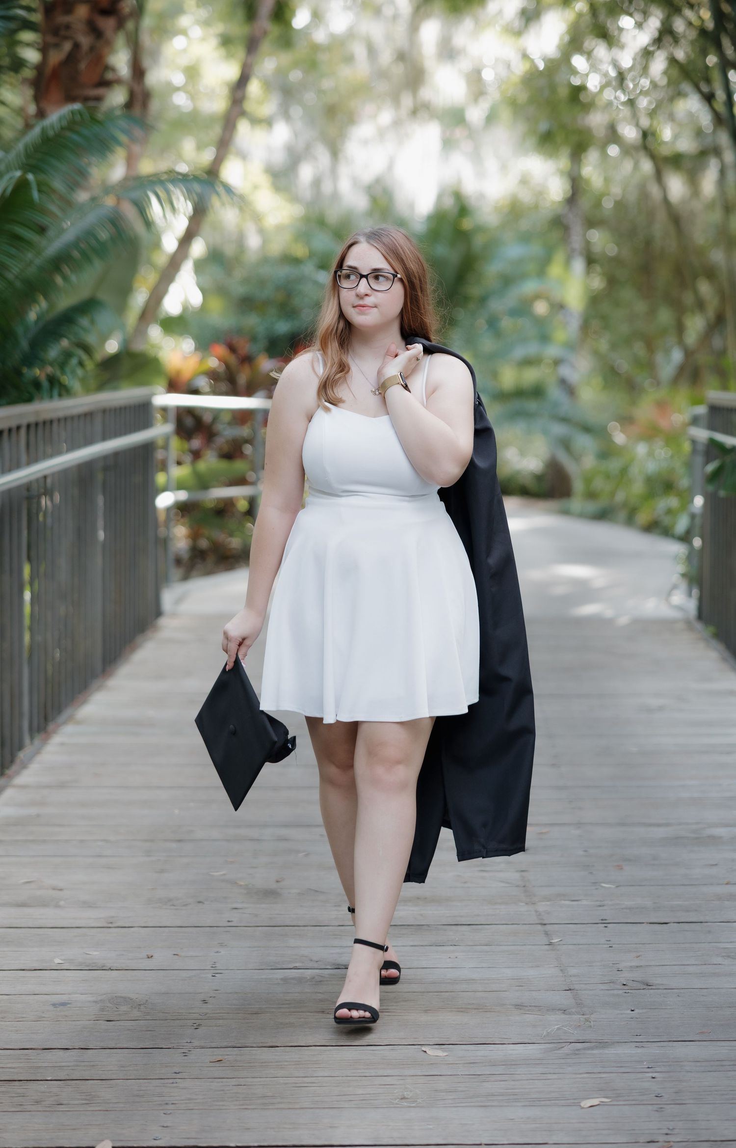 Graduate walks along wooden boardwalk carrying graduation cap and gown.