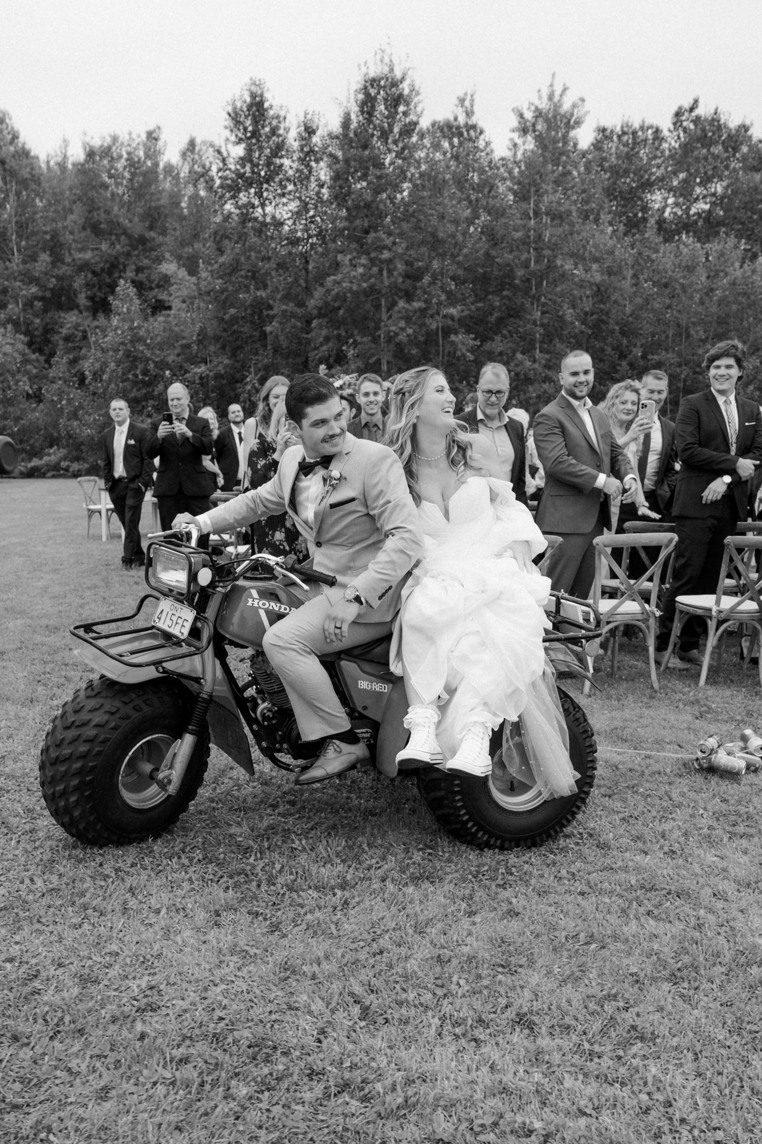 Wedding couple rides off on mini dirt bike as guests cheer in black and white photo.