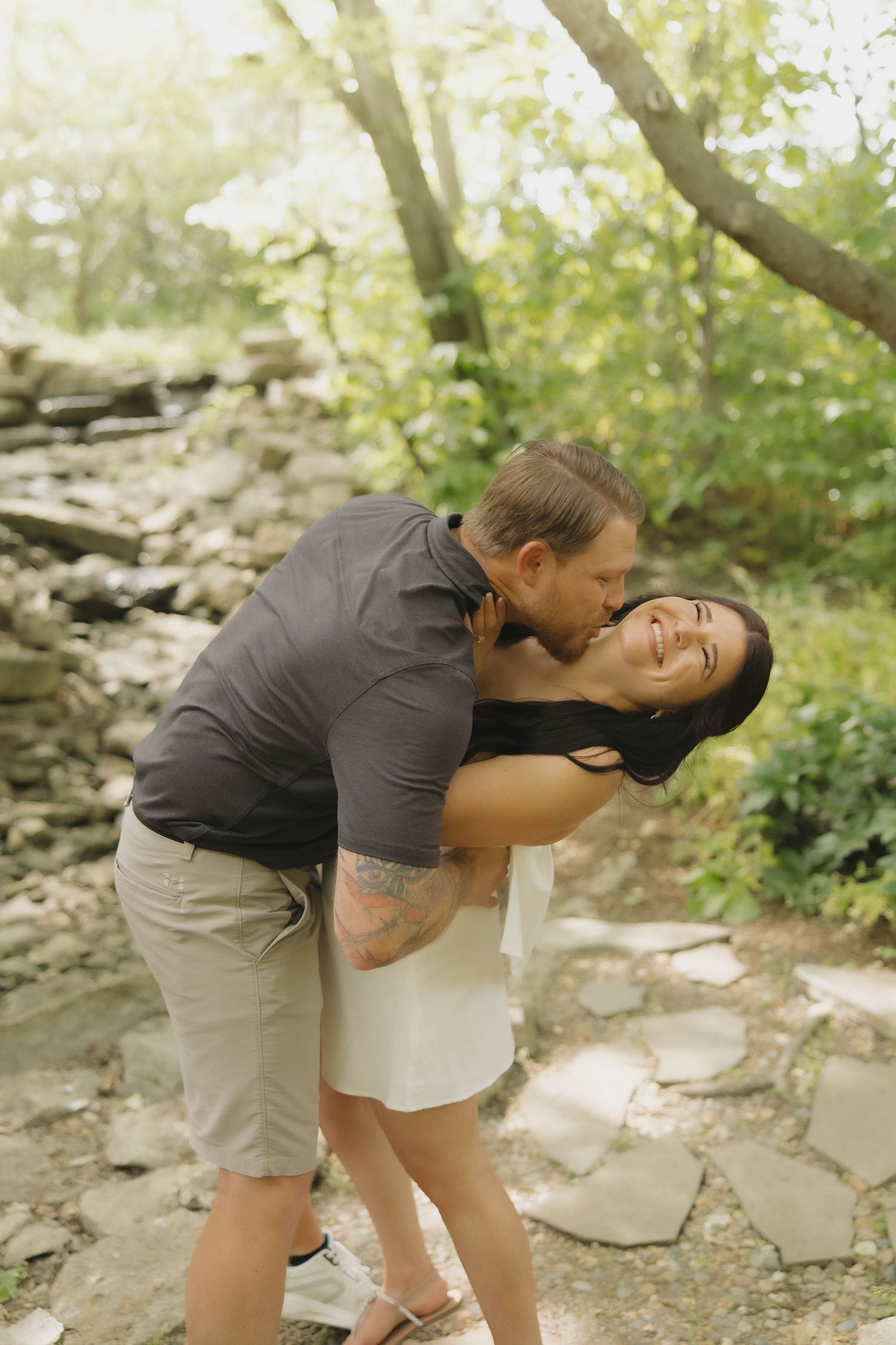 A romantic couple shares a playful moment on a stone path surrounded by lush greenery and natural sunlight.