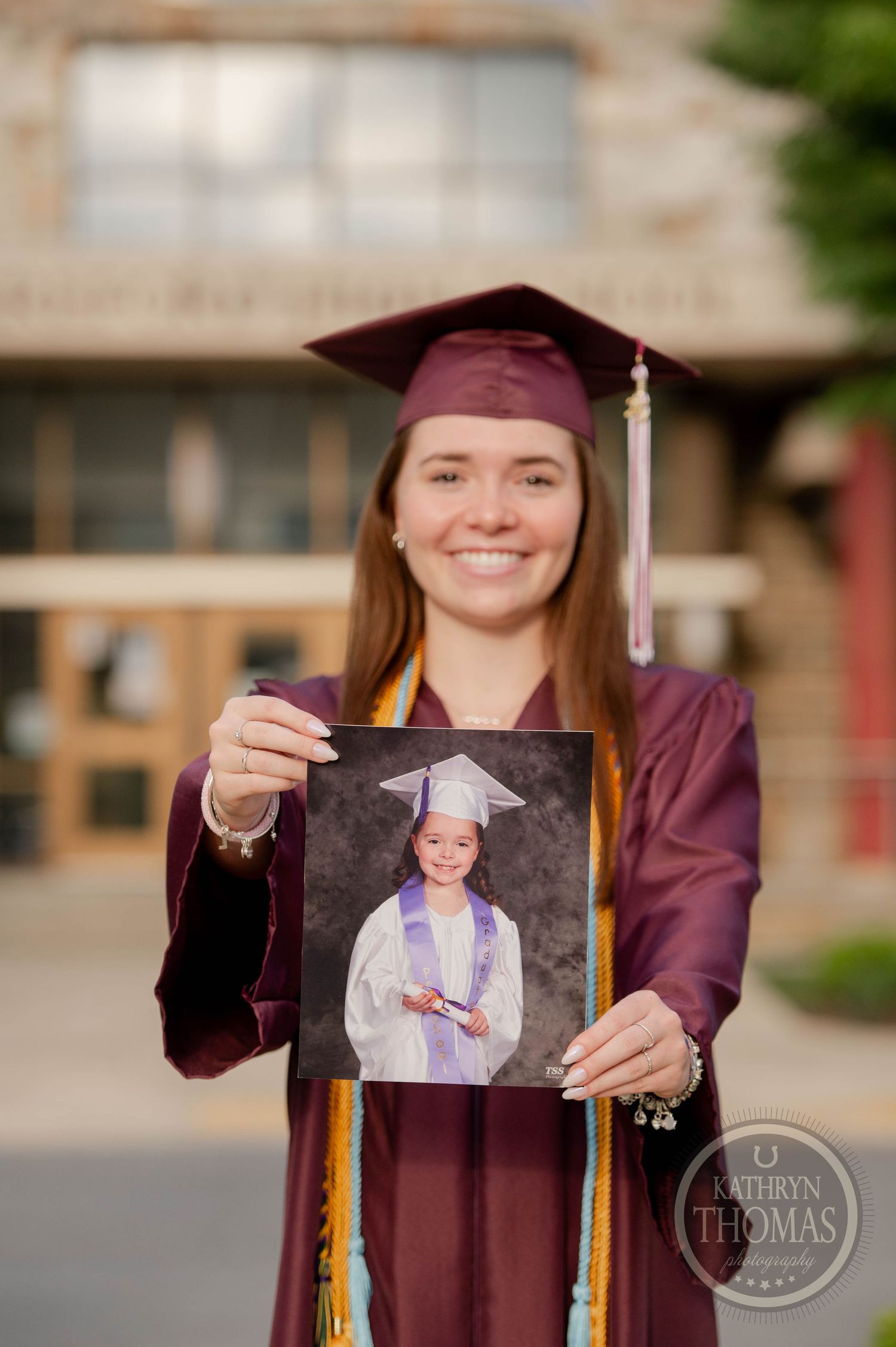 Graduate in maroon cap and gown holds up a framed graduation photo while standing outdoors.