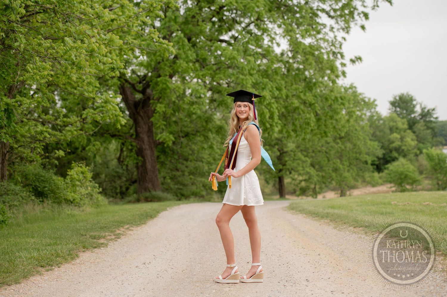 Graduate in white dress holding bouquet of sunflowers poses on dirt path surrounded by green trees on sunny day.