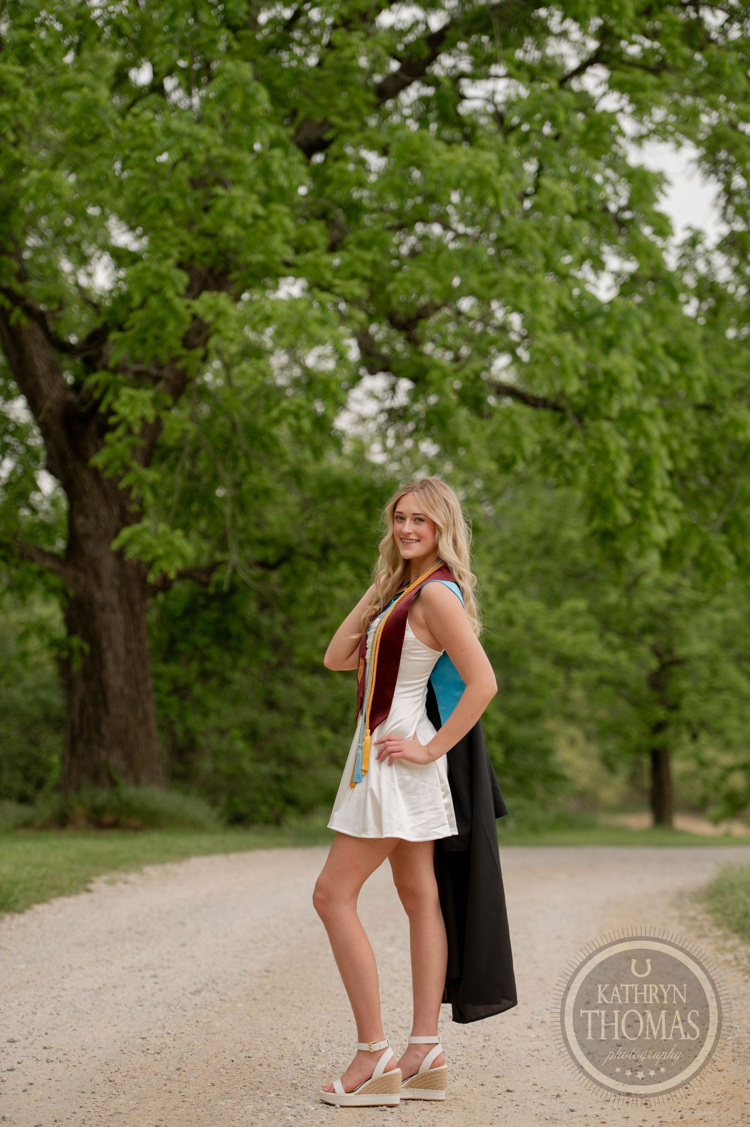 A graduate stands on a dirt path wearing a white dress and holding graduation attire, surrounded by green trees in summer.