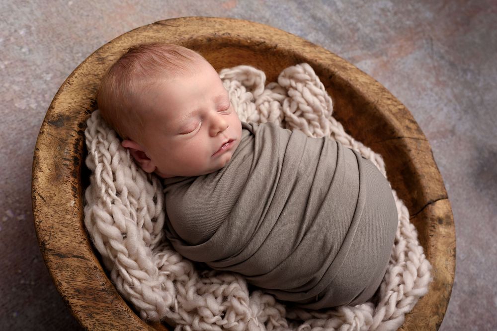 Peaceful newborn swaddled in brown fabric nestled in wooden bowl with knit blanket.