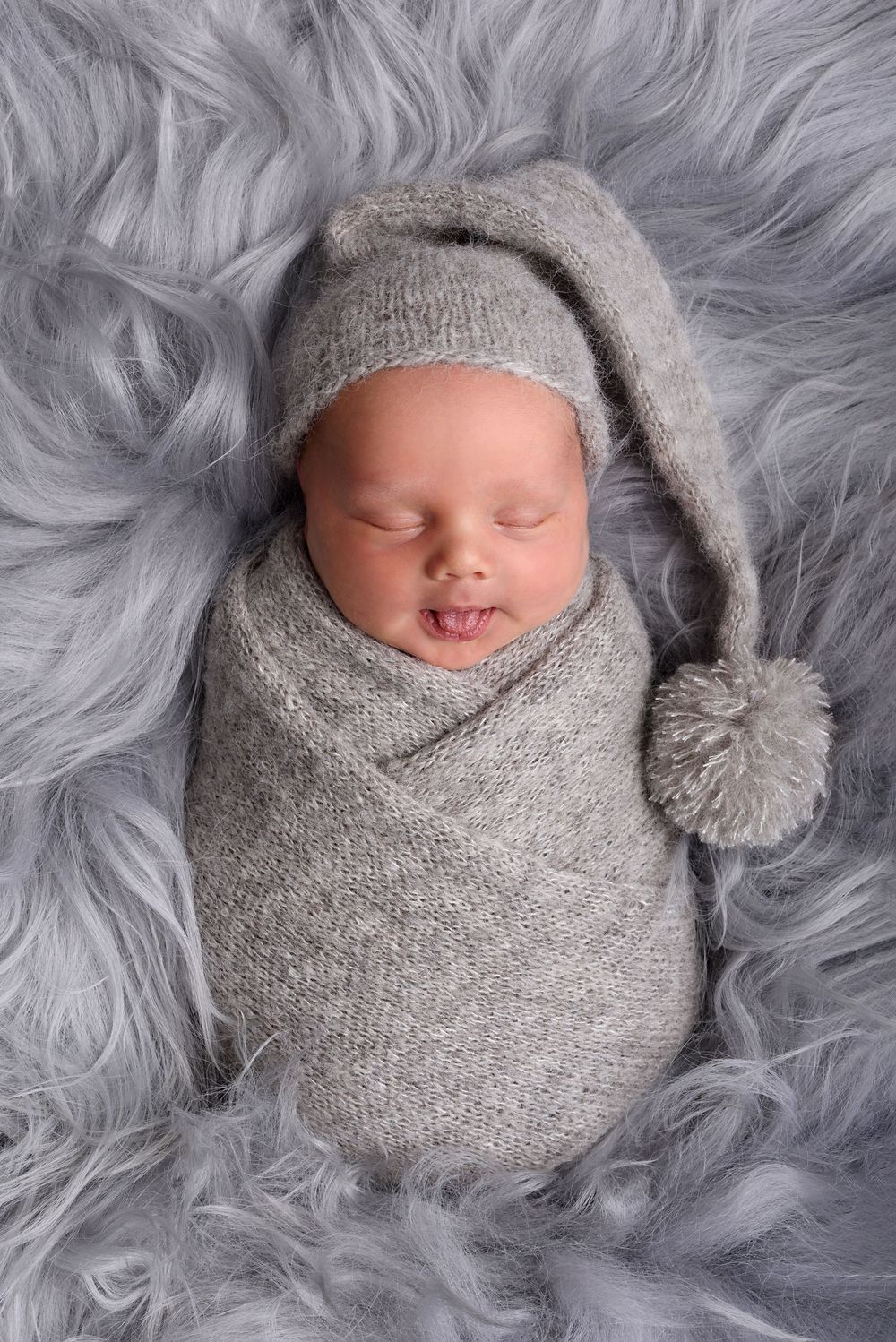 Sleeping newborn wrapped in gray knit with pom pom hat on furry backdrop.