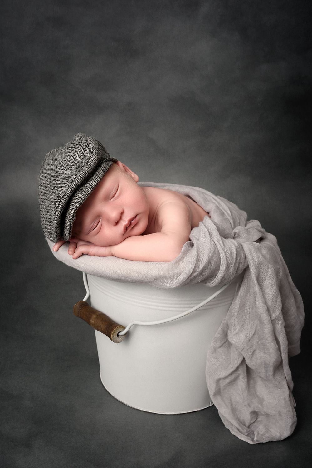 Sleeping newborn wearing gray cap rests in white bucket with draped fabric against dark background.