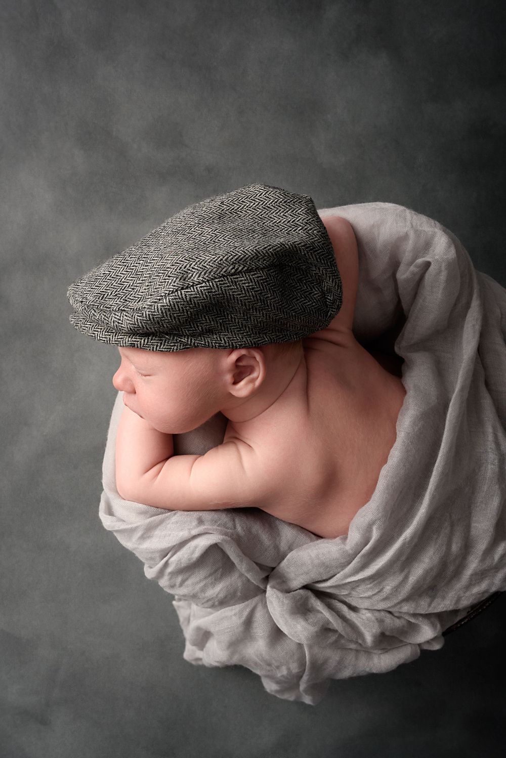 Peaceful newborn wearing newsboy cap wrapped in gray fabric against moody background.