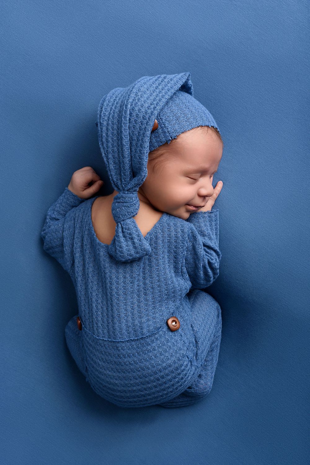 Sleeping baby in textured blue outfit with matching bonnet against blue background.