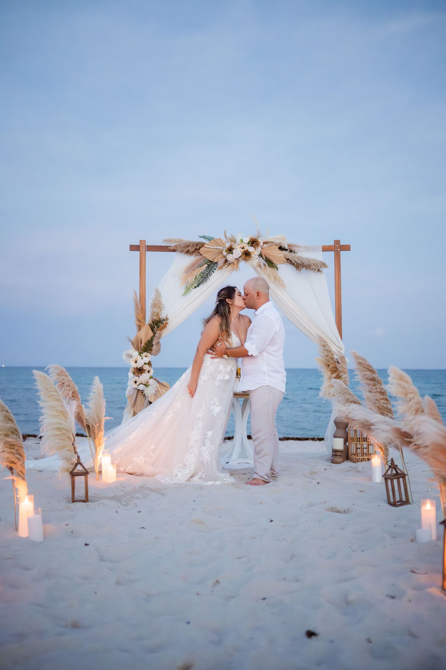 A romantic beach wedding setup with wooden arches decorated with pampas grass and candles on the sand at sunset.