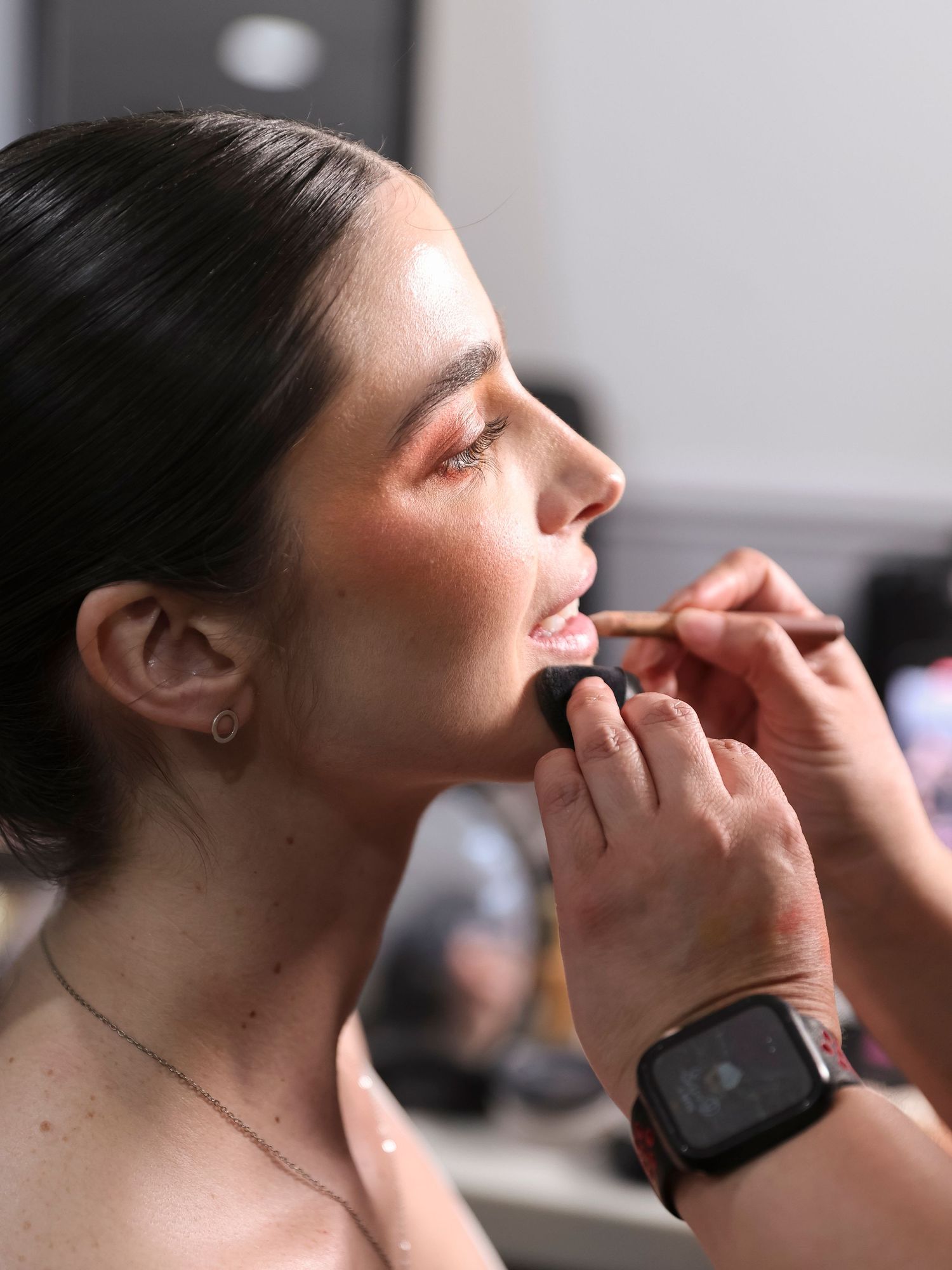 An artist applies lip color with a makeup brush while wearing a smartwatch during a professional beauty session.
