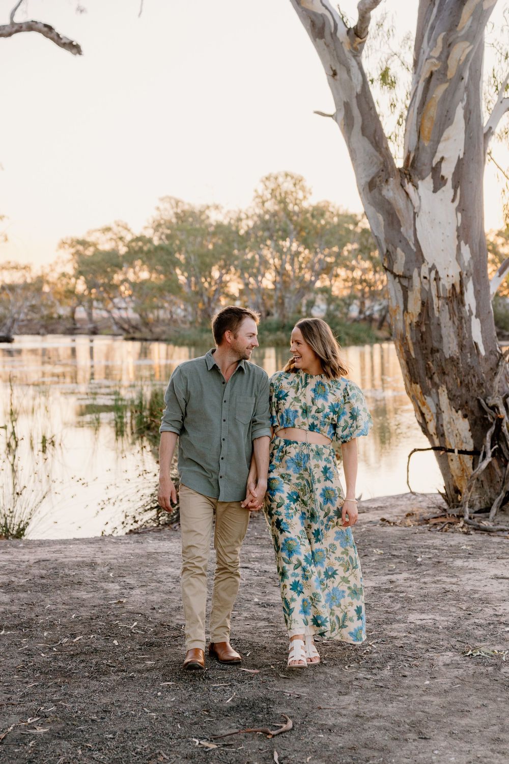A couple walks hand in hand along a riverbank at sunset with a large eucalyptus tree in the background.