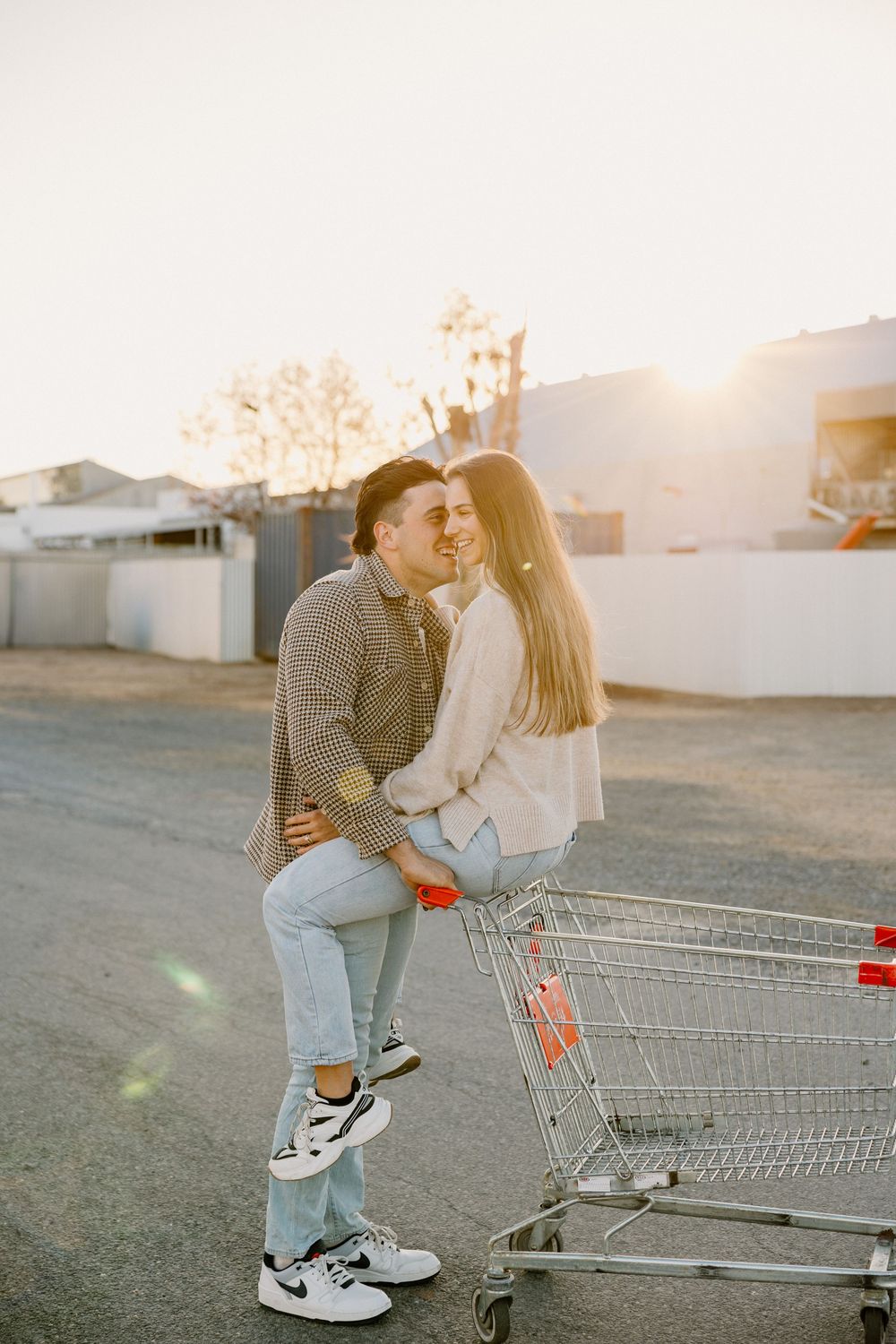 A playful couple shares an affectionate moment by a shopping cart in a parking lot during golden hour sunset.