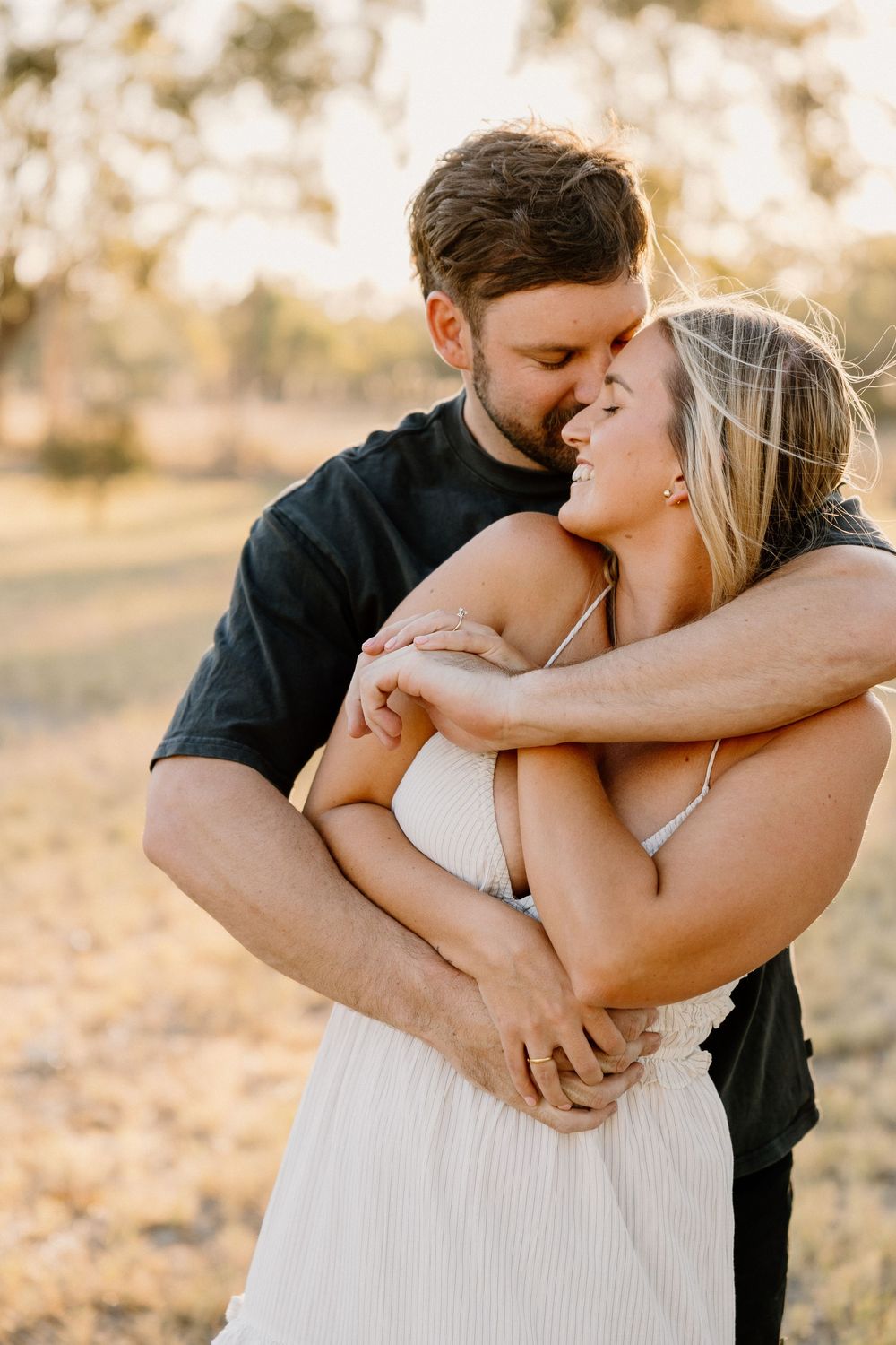 Romantic couple embraces during sunset photoshoot in a field, sharing intimate moment while dressed in black and white.