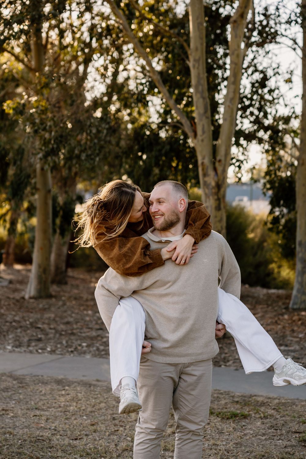 A couple shares a joyful embrace in a casual outdoor setting with trees during golden hour.
