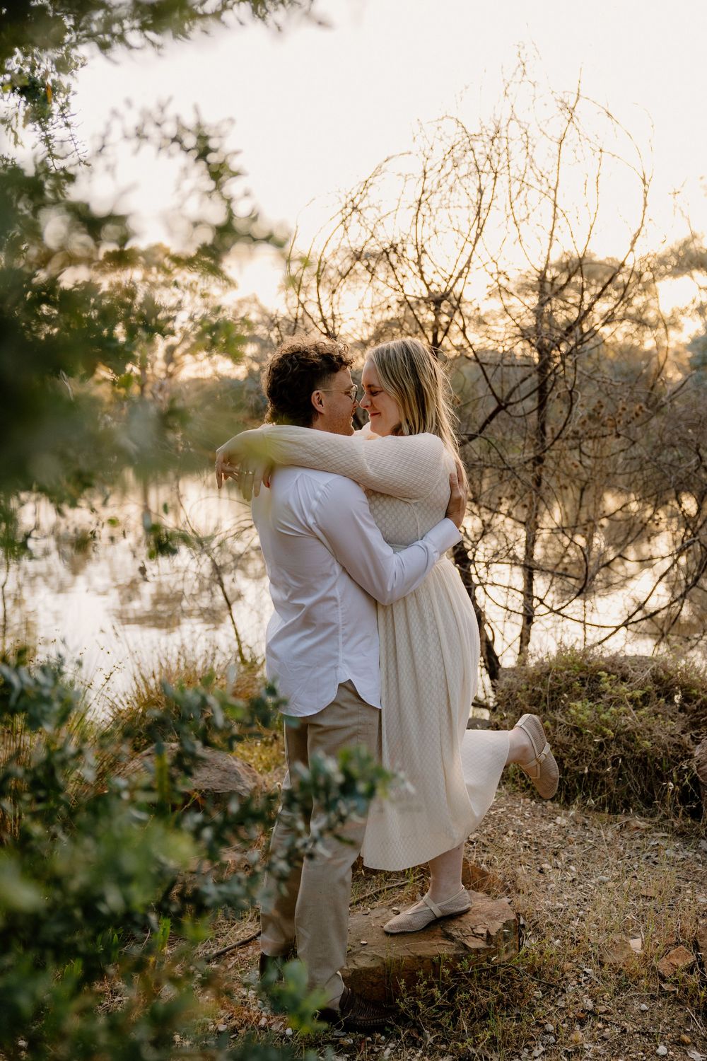 Couple embraces in romantic sunset moment by lake with golden light filtering through trees and branches.