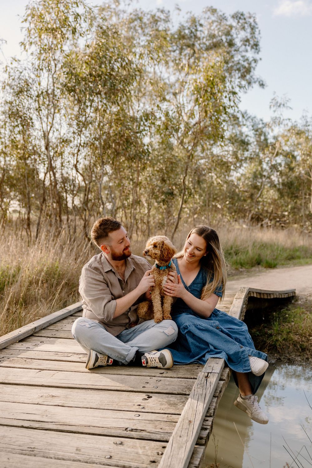 Couple sits with their golden retriever puppy on wooden dock by lake surrounded by eucalyptus trees at sunset.