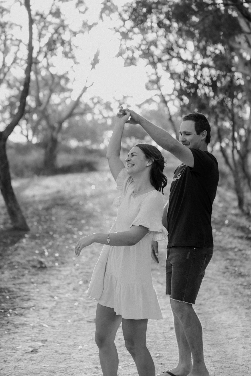 A couple dances together on a forest path in black and white, with the woman twirling in a white summer dress.