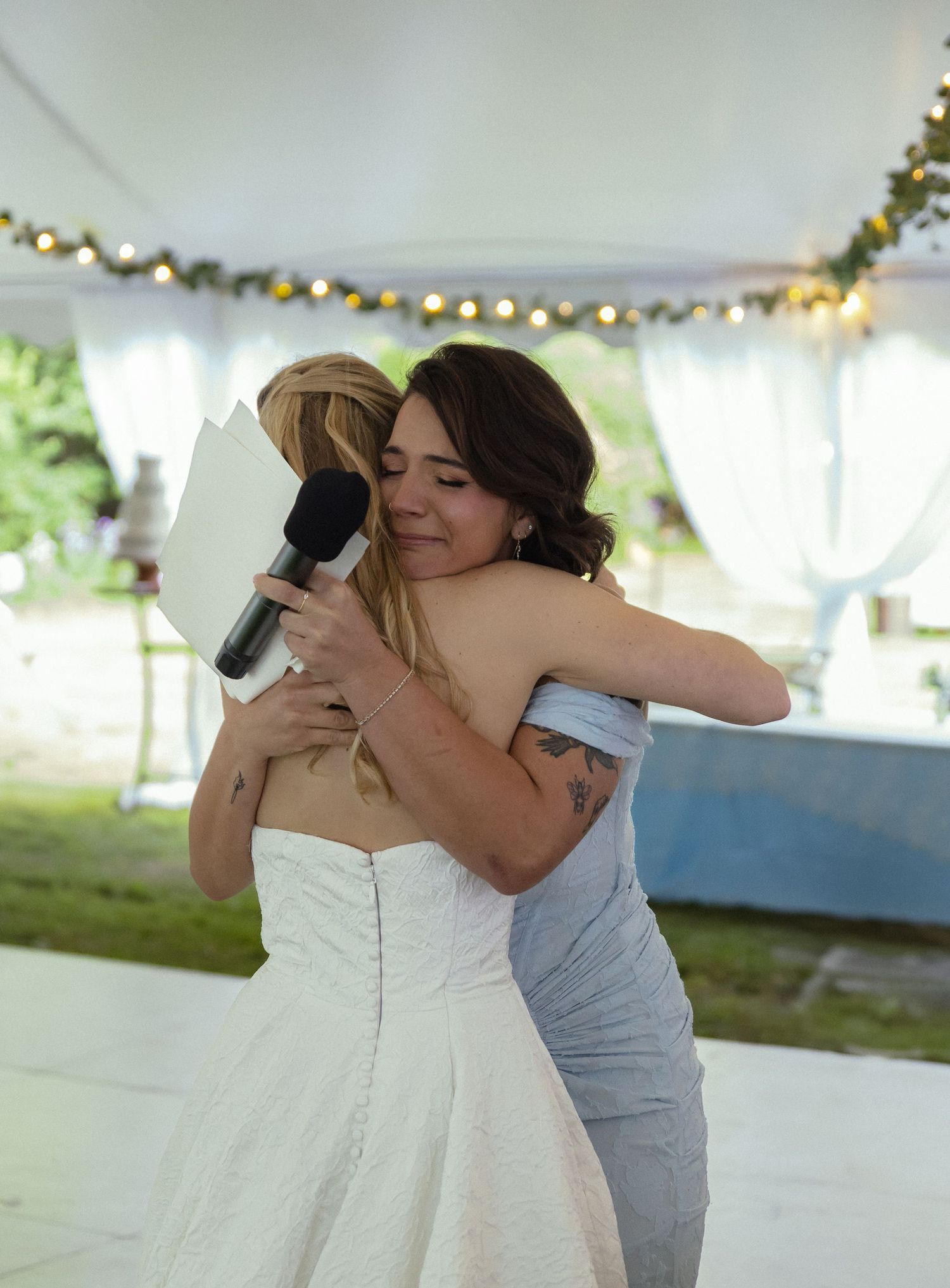 Emotional embrace during a wedding celebration under a white tent with twinkle lights.