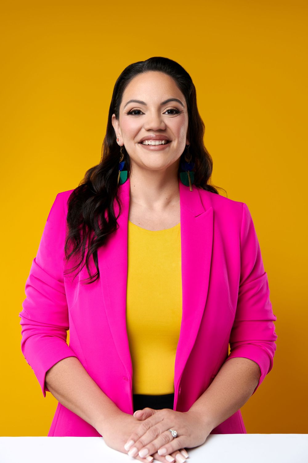 Professional portrait of someone wearing a vibrant hot pink blazer and yellow top against a bright yellow background.