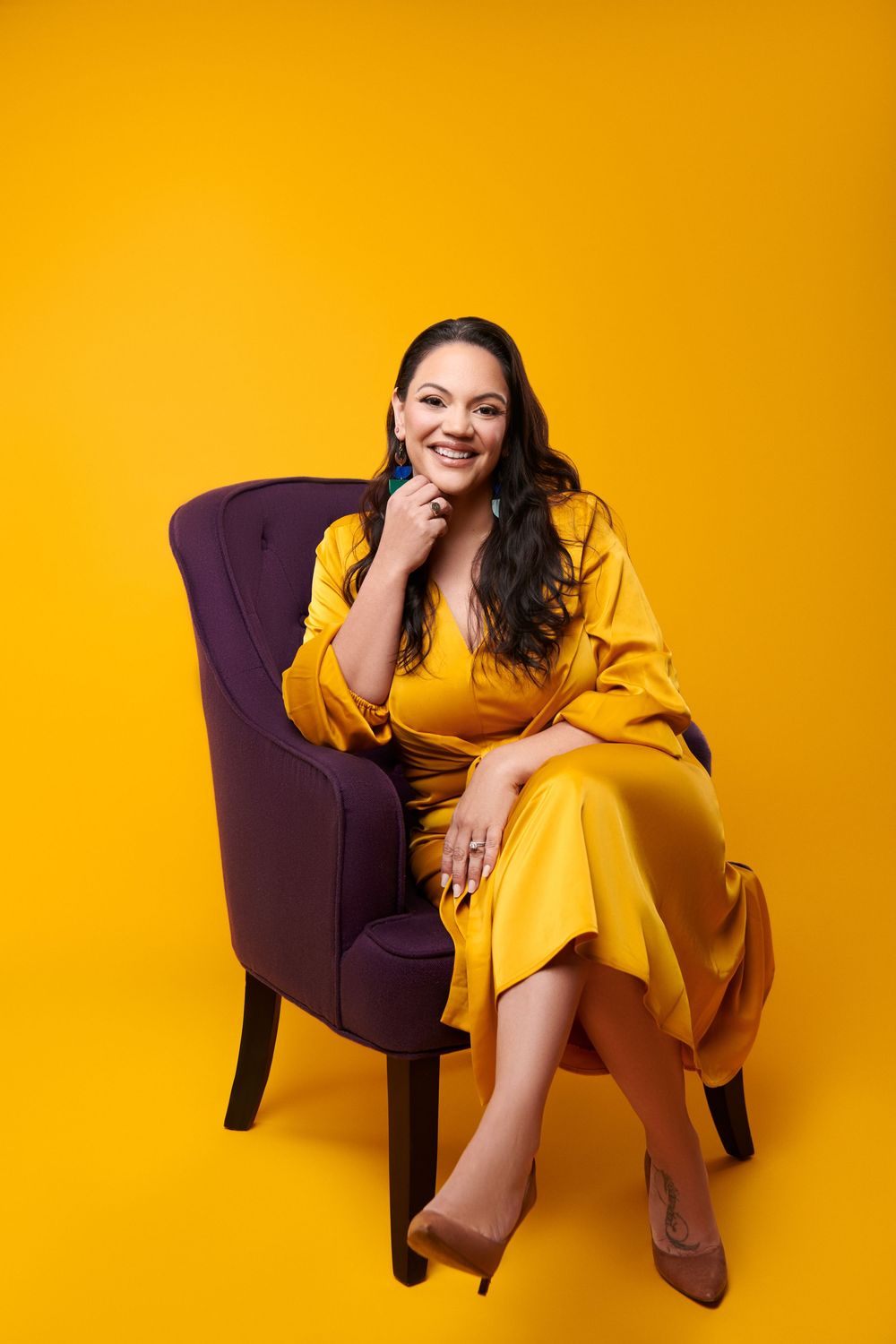 A person in a yellow dress sits elegantly on a purple armchair against a vibrant yellow background.