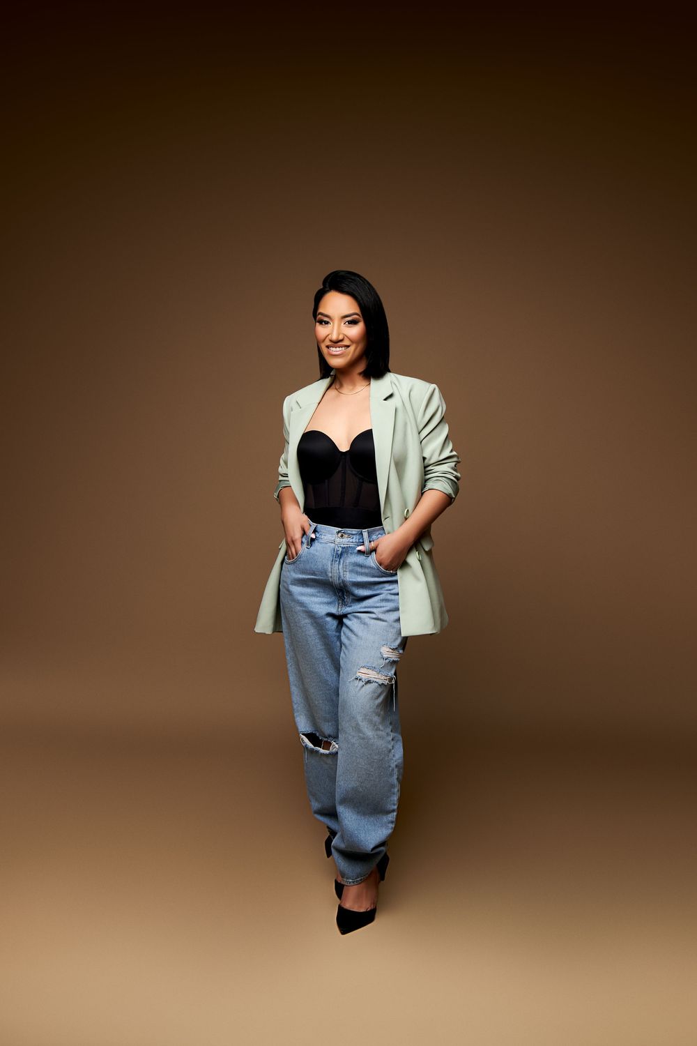 Fashion model wears oversized denim overalls paired with a mint green blazer against brown studio backdrop.