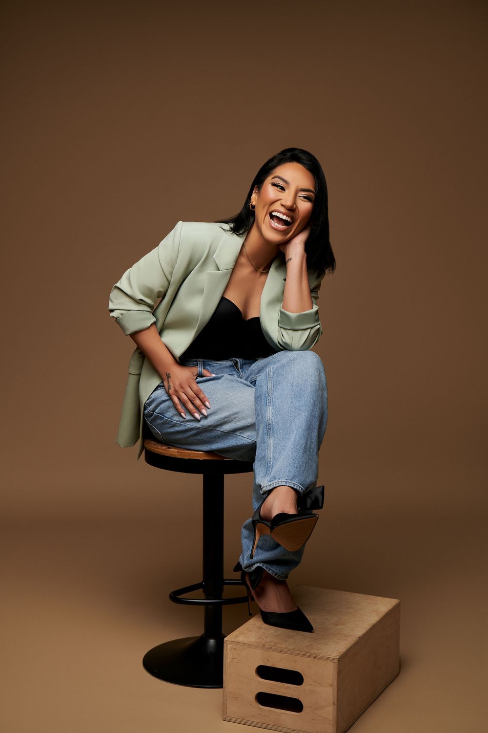 A person in a mint green blazer and jeans sits casually on a black bar stool, laughing joyfully against a brown backdrop.