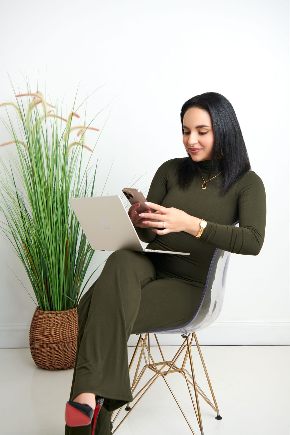 Person in olive green dress sits in modern white chair with woven basket and plant, using mobile devices.